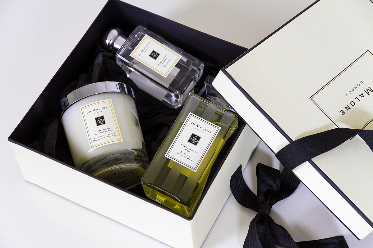 Jo Malone London, living in a scented home, open cream signature gift box and black ribbon, inside the box are Jo Malone products, a scented candle, a bath oil and a cologne