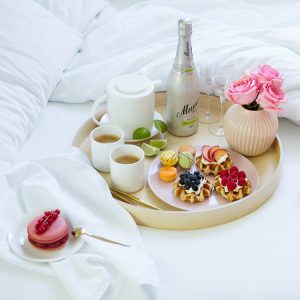 breakfast in bed tray waffles macarons fruits coffee bottle of champagne vase pink flowers white linen